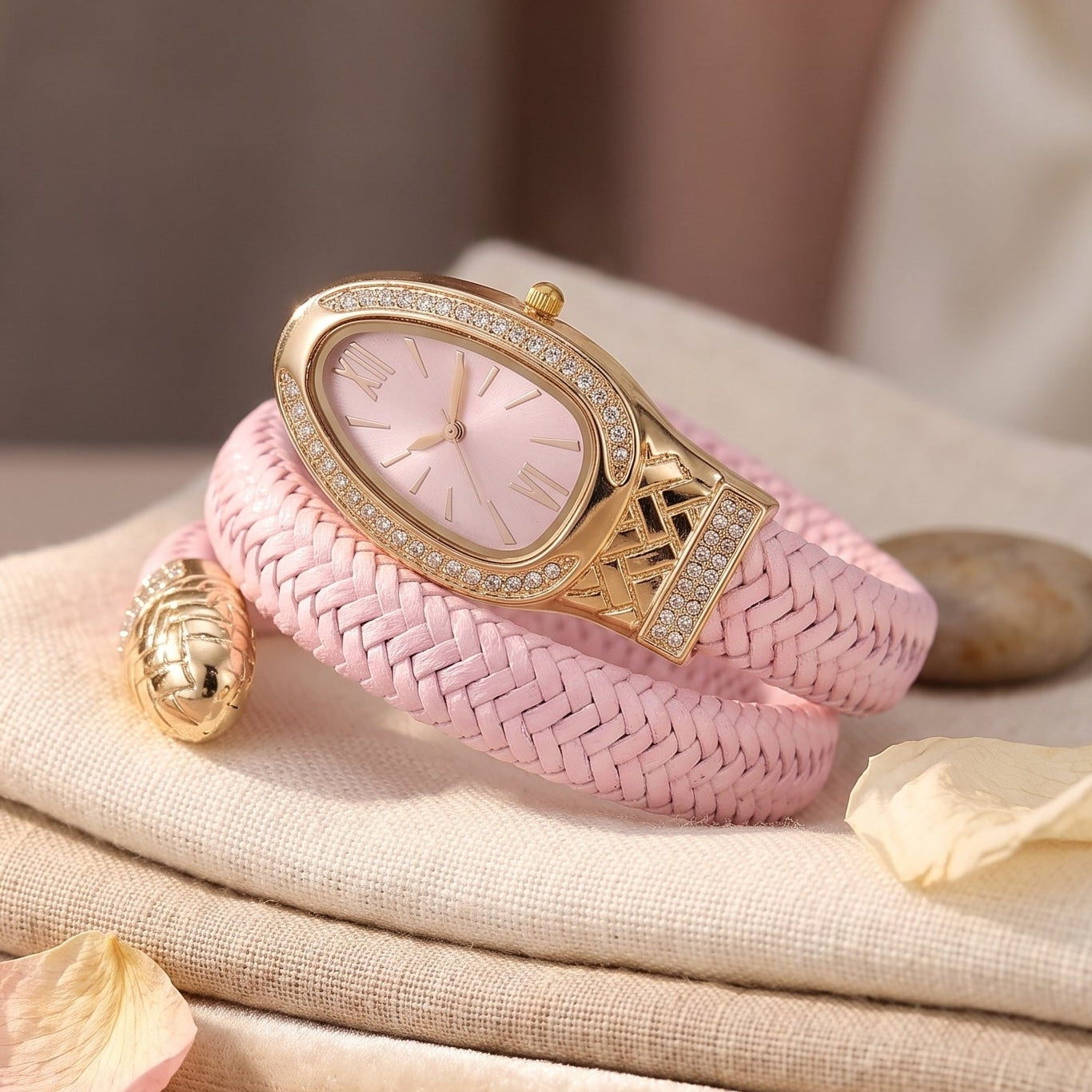 A pink braided woven watch with a gold-tone case and crystal-embellished bezel, styled on folded beige linen with a small gold shell accessory and soft petals.