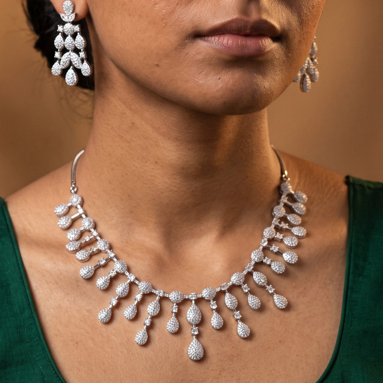 A woman wearing a silver-toned teardrop pendant necklace and matching drop earrings with clear stones, shown against a warm brown studio background.