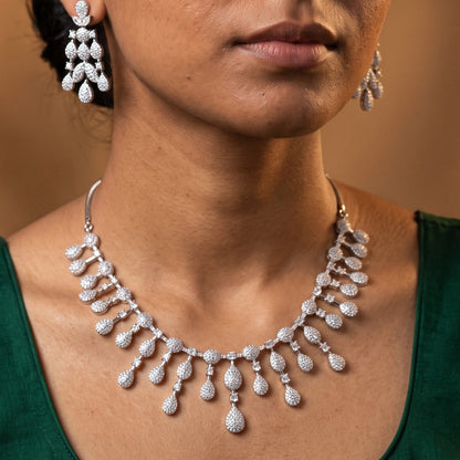 A woman wearing a silver-toned teardrop pendant necklace and matching drop earrings with clear stones, shown against a warm brown studio background.
