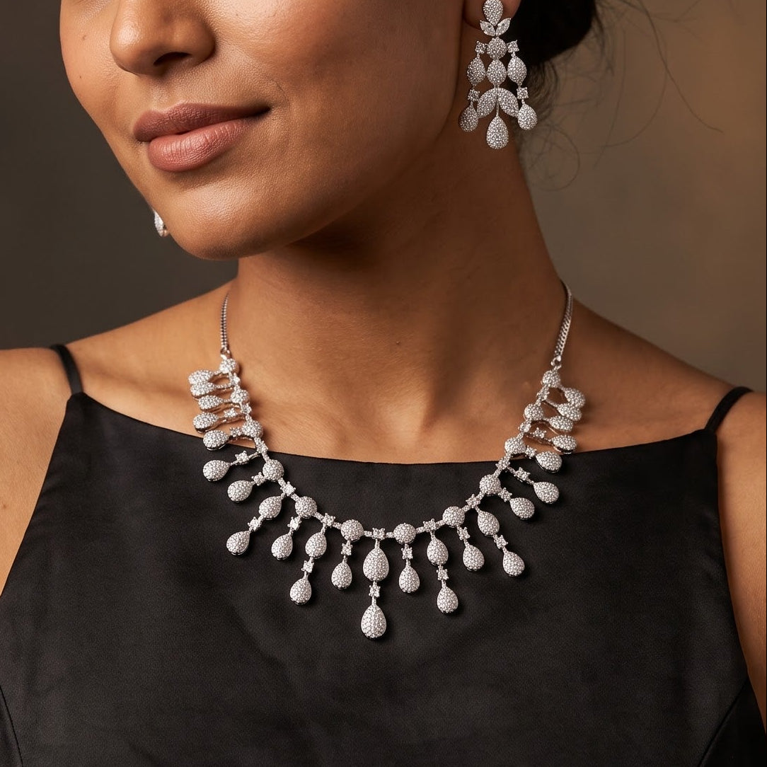 A woman with dark hair tied back wears a silver-toned statement necklace and matching drop earrings with clear teardrop-shaped stones, paired with a black satin top against a dark brown studio background.