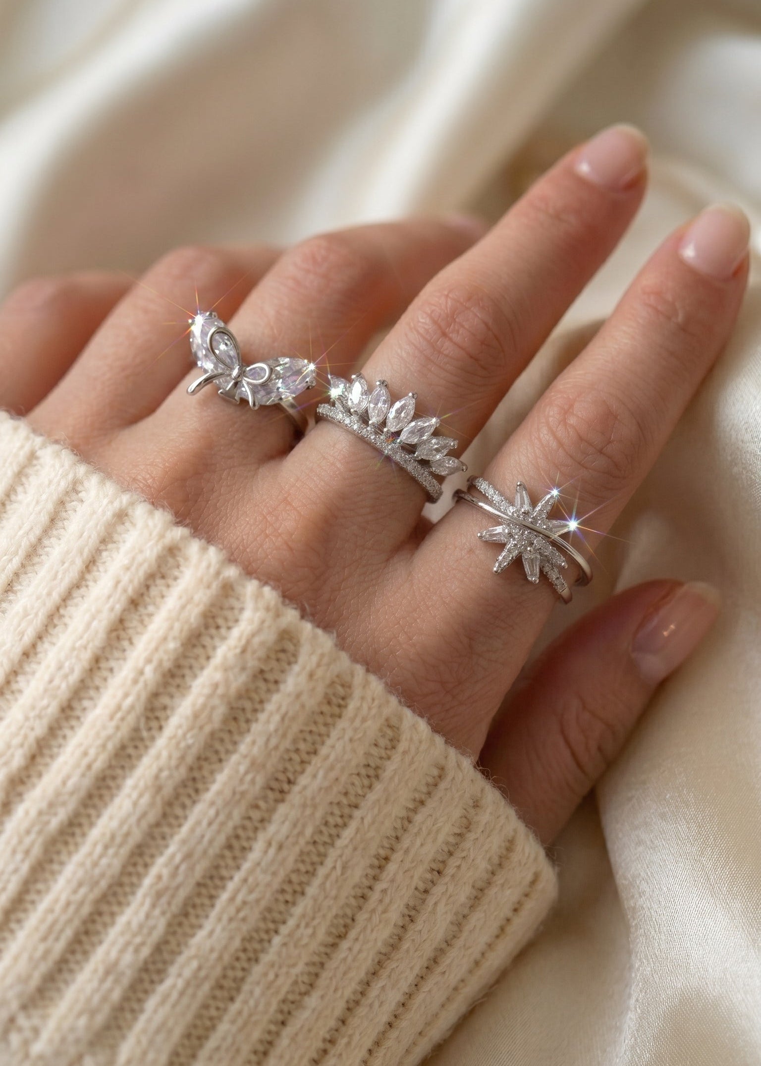 A close-up of a hand wearing three silver rings with clear crystal details, styled with a cream-colored ribbed knit sweater against a soft cream fabric background.